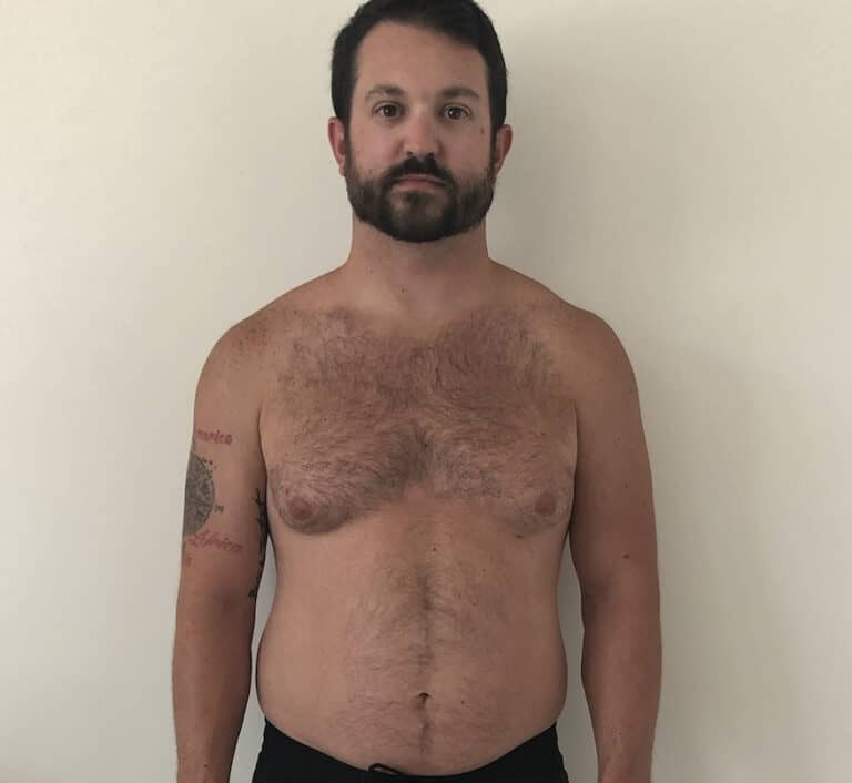 A man with short dark hair and a beard stands shirtless against a plain white wall, showcasing his fitness in black shorts. He has a tattoo on his left upper arm and the confident look of an online fitness coach.
